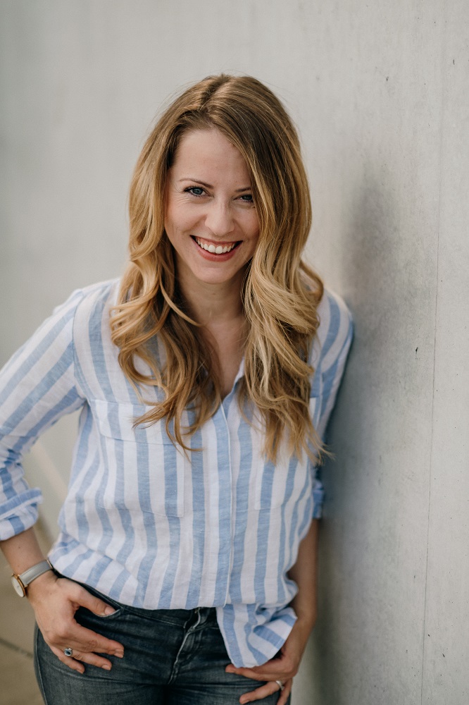 Das Bild zeigt die Expertin für Zeitmanagement Julia Leifheit, wie diese lachend und an einer Wand lehnend in die Kamera blickt. Sie trägt eine blau-weiß gestreifte Bluse.