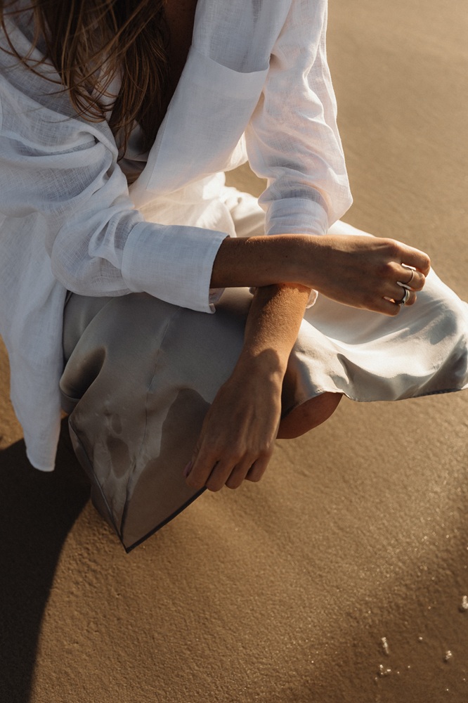 Frau in beigem Kleid und weißem Hemd hockt im Sand, Hände überkreuzt